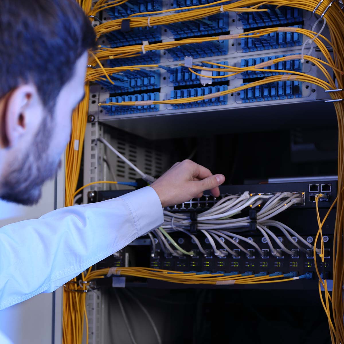 Das Bild zeigt einen männlichen IT-Ingenieur, der in einem Serverraum arbeitet und für die Netzwerksicherheit sorgt. Er überprüft sorgfältig die angeschlossenen Kabel an einem Server, was auf eine präzise und verantwortungsvolle Arbeitsweise in der IT-Infrastruktur hinweist. Der Raum ist mit zahlreichen Servern und technischen Geräten ausgestattet, die eine professionelle Arbeitsatmosphäre vermitteln.