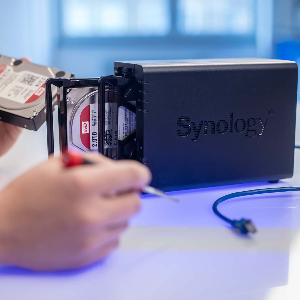 Das Bild zeigt einen Mitarbeiter, der eine NAS-HDD (Network-Attached Storage Hard Drive) austauscht. Der Mitarbeiter arbeitet sorgfältig und präzise, um die Festplatte zu entfernen und durch eine neue zu ersetzen. Die Szene vermittelt einen technischen Arbeitsprozess in einer professionellen IT-Umgebung, bei dem auf Datenintegrität und Netzwerkoptimierung geachtet wird.