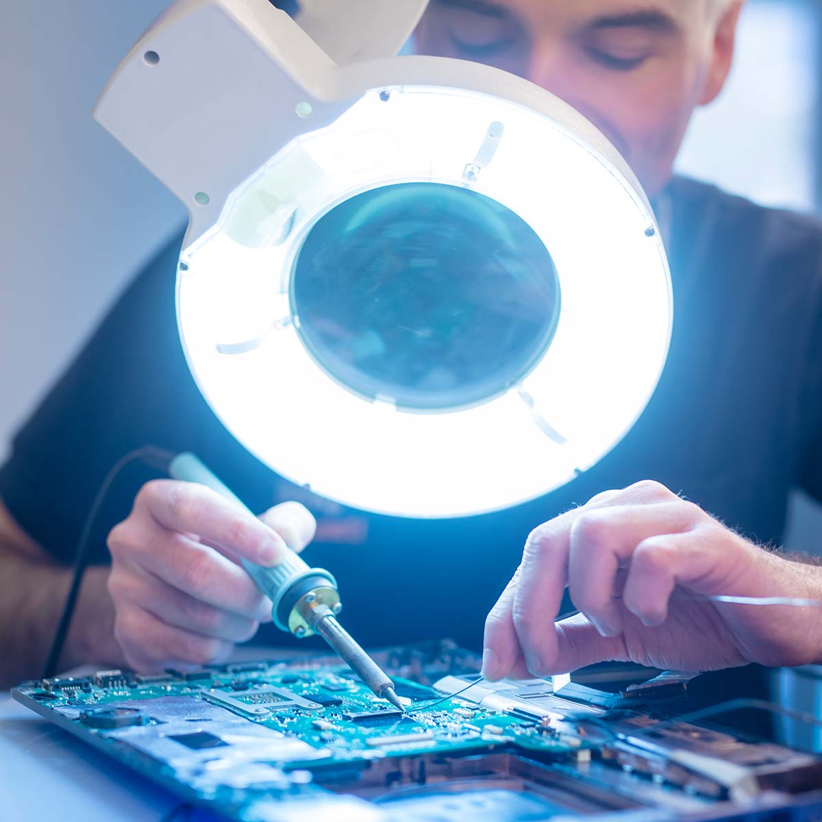 Das Bild zeigt einen Mitarbeiter, der Ersatzteile einlötet. Mit präziser Handarbeit arbeitet er an einer technischen Komponente, um diese zu reparieren oder zu optimieren. Die Szene vermittelt eine detailorientierte und technische Arbeitsumgebung, in der Fachwissen und Sorgfalt erforderlich sind.