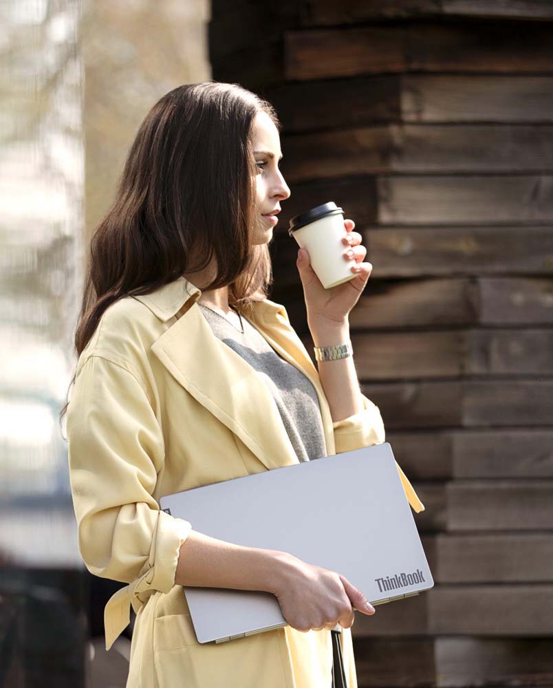 Lifestyle Foto Lenovo ThinkBook Eine Frau in einem gelben Blazer trinkt entspannt Kaffee und hält in ihrer rechten Hand ein Lenovo ThinkBook. Die Szene strahlt Professionalität und Eleganz aus, während sie das ThinkBook als Teil ihres täglichen Arbeitsalltags nutzt.